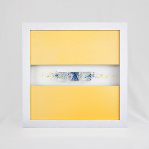 May include: A framed artwork with a yellow background and a white mat. The artwork features three small blue and white squares with a blue and white design inside each square. The squares are arranged in a row and are surrounded by yellow and white paint splatters.