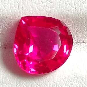 Può includere: Una gemma sfaccettata a forma di pera di un colore rosa acceso e saturo. La pietra presenta molteplici sfaccettature geometriche che riflettono la luce, creando un aspetto brillante e scintillante su tutta la superficie lucida e traslucida.