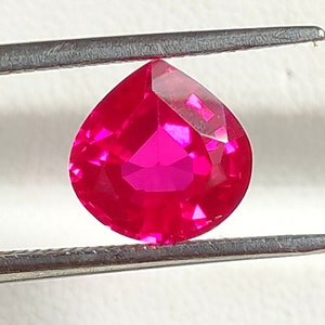 Può includere: Una gemma a forma di pera di un vibrante rosso fucsia con una superficie sfaccettata che riflette la luce. La gemma è tenuta tra le due punte metalliche di una pinzetta color argento, mettendo in risalto il suo colore chiaro e saturo.