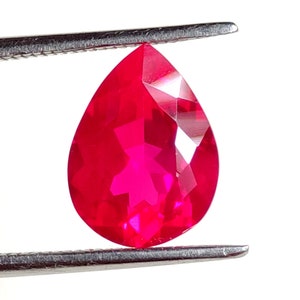 Könnte beinhalten: Ein birnenförmiger, facettierter Edelstein in einer leuchtenden, gesättigten roten Farbe. Der Stein wird von einer silberfarbenen Metallpinzette gehalten und zeigt seine klare, reflektierende Oberfläche sowie präzise geometrische Schliffe.