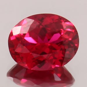 May include: A faceted, oval-shaped ruby gemstone. The gem is a deep, vibrant red color with multiple facets that reflect light. The ruby is set against a light background, with its reflection visible below.