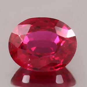 May include: A vibrant, oval-cut gemstone with a deep, saturated red color and multiple facets that create a brilliant, reflective surface. The stone displays a clear, transparent quality with sharp edges and a polished finish, showing a distinct reflection.
