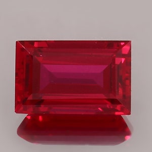 May include: A vibrant, deep red gemstone cut into a rectangular emerald shape. The stone features sharp, clean edges and a faceted surface that catches the light, creating a clear reflection on the surface below it, showcasing its intense color.