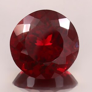 Pode incluir: Uma pedra preciosa redonda, facetada, de cor vermelha profunda. A gema é altamente polida, refletindo a luz e criando um brilho brilhante. A pedra preciosa está sobre um fundo branco, com seu reflexo visível abaixo.