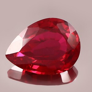 Könnte beinhalten: Ein birnenförmiger, facettierter Edelstein in einer tiefen, leuchtend roten Farbe. Der Stein hat eine polierte Oberfläche mit mehreren geometrischen Facetten, die das Licht reflektieren und ein brillantes Aussehen vor einem neutralen Hintergrund erzeugen.