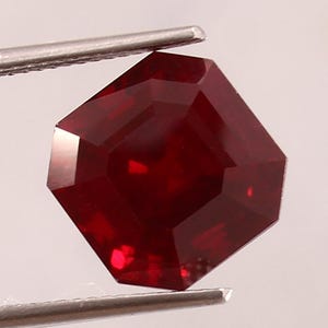 May include: A deep red, faceted gemstone with an octagonal shape held by metal tweezers. The stone displays a rich, saturated crimson hue with sharp, geometric edges and a polished surface that reflects light across its multiple flat facets.