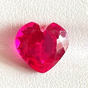 May include: A vibrant, heart-shaped gemstone with a deep, saturated red color and a faceted surface that reflects light. The stone features a classic heart cut with a pointed base and a cleft at the top, showcasing a brilliant, polished finish.