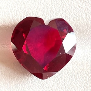 May include: A heart-shaped gemstone with a deep, vibrant red color and multiple faceted surfaces that reflect light. The stone features a classic heart silhouette with a pointed base and a rounded top indentation, showcasing a clear, polished finish.