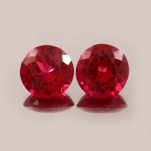 Könnte beinhalten: Zwei runde, facettierte, leuchtend rote Edelsteine, die nebeneinander liegen. Jeder Stein weist einen Brillantschliff mit zahlreichen geometrischen Facetten auf, die das Licht reflektieren und eine tiefrote Farbe sowie eine klare Optik erzeugen.