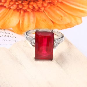 May include: A silver ring featuring a large, rectangular, deep red gemstone set in the center. The band is accented with small, clear, round stones along the shoulders, providing a sparkling contrast to the vibrant red center stone.