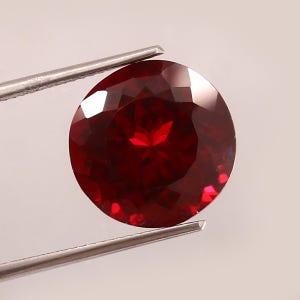May include: A round, deep red gemstone with multiple facets is held by silver tweezers. The gem reflects light, highlighting its rich colour. The background is white. This could be a ruby or garnet, a gemstone often used in jewellery.