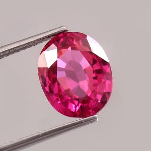 Può includere: Una gemma dal taglio ovale di un vibrante colore rosso rosato, tenuta da una pinzetta in metallo color argento. La pietra presenta una superficie sfaccettata che riflette la luce, creando motivi geometrici distinti sulla superficie lucida.