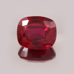 May include: A cushion-cut ruby gemstone with a deep, saturated red colour and a faceted surface that reflects light. The stone displays a clear, transparent quality with internal depth and a distinct reflection visible directly beneath it.