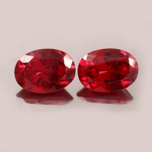 May include: Two oval-cut, vibrant red gemstones with multiple facets that reflect light. The stones are positioned side-by-side, displaying a deep, saturated crimson color and a polished, reflective surface with sharp, geometric edges.