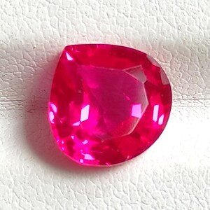 Può includere: Una gemma vibrante a forma di pera con un colore rosa fucsia profondo e saturo. La pietra presenta una superficie sfaccettata che riflette la luce, creando molteplici riflessi interni e ombre su tutto il suo corpo lucido e brillante.