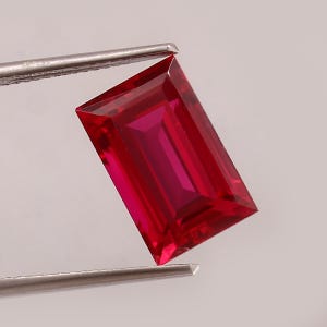 May include: A vibrant, rectangular-cut red gemstone with a faceted surface that reflects light. The stone is held by a pair of silver-toned metal tweezers, showcasing its deep crimson color, sharp geometric edges, and clear, transparent clarity.