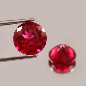 Könnte beinhalten: Ein runder, facettierter, leuchtend roter Edelstein, der von einer silberfarbenen Metallpinzette gehalten wird. Ein zweiter, identischer runder, facettierter roter Edelstein befindet sich leicht unscharf rechts und zeigt eine klare Spiegelung.