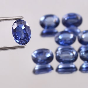 Op de afbeelding: Een ovaalvormige, diepblauwe edelsteen wordt vastgehouden door een pincet, met verschillende andere vergelijkbare edelstenen verspreid op de achtergrond. De edelstenen hebben een gepolijst, reflecterend oppervlak en de belichting creëert highlights en schaduwen.