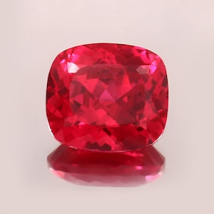 Peut inclure: Une pierre précieuse taillée en coussin, à facettes, d'un rouge vif. La gemme est très réfléchissante, avec de multiples facettes captant la lumière. La pierre est présentée sur une surface réfléchissante, créant un effet miroir.