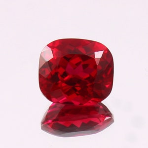 May include: A cushion-cut loose gemstone with a deep, vibrant red color and multiple facets that reflect light. The stone has a square shape with rounded corners and exhibits a high level of clarity, showing a clear reflection on the surface below.