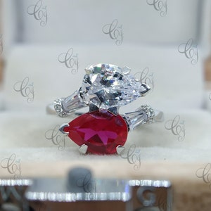 May include: A silver ring with two pear-shaped gemstones. One is clear, the other is a deep red. The ring has a distinctive design, with smaller, rectangular stones set on either side of the larger stones.