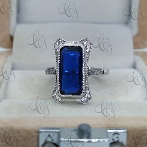 May include: A silver ring with a large, rectangular, deep blue gemstone at the center. The ring features an ornate, filigree design around the gemstone, accented with small, clear stones. The ring is set in a velvet-lined box.
