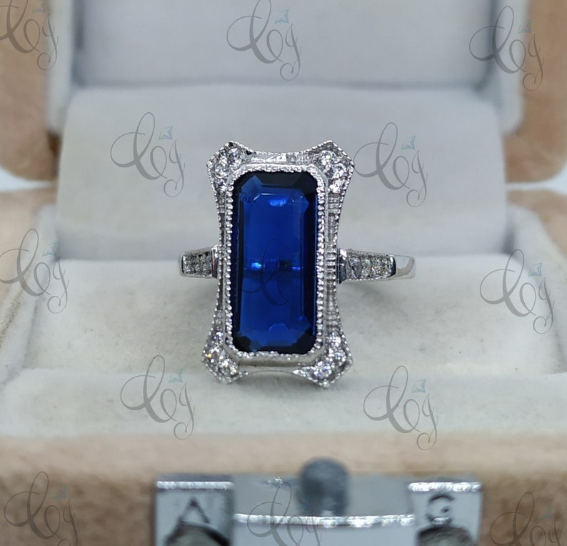 May include: A silver ring with a large, rectangular, deep blue gemstone at the center. The ring features an ornate, filigree design around the gemstone, accented with small, clear stones. The ring is set in a velvet-lined box.