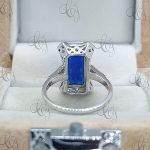 May include: A silver ring with a rectangular, deep blue gemstone. The ring features an ornate, openwork design around the stone and along the band. The ring is set in a light-colored box.