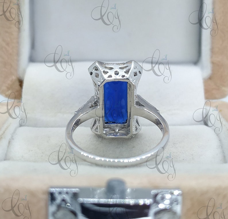 May include: A silver ring with a rectangular, deep blue gemstone. The ring features an ornate, openwork design around the stone and along the band. The ring is set in a light-colored box.