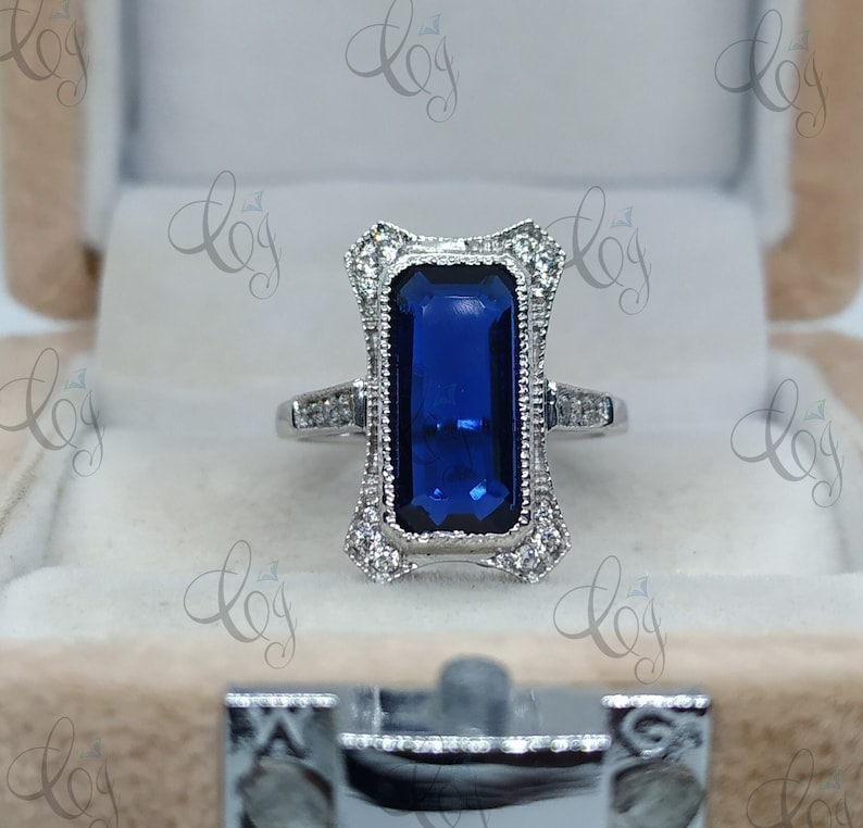 May include: A silver ring with a rectangular, deep blue gemstone. The ring features a decorative, ornate setting with small, clear accent stones. The ring is displayed in a beige jewelry box, showcasing its elegant design.