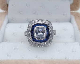 May include: A silver ring with a square-shaped center stone surrounded by a double halo of smaller stones. The inner halo is blue and the outer halo is clear.