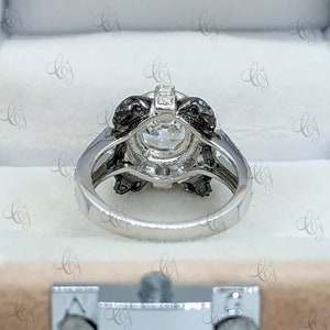 May include: A silver engagement ring with a large, round, clear center stone surrounded by a black halo setting. The ring is in a white box.