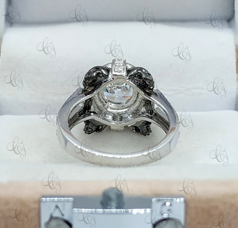 May include: A silver engagement ring with a large, round, clear center stone surrounded by a black halo setting. The ring is in a white box.