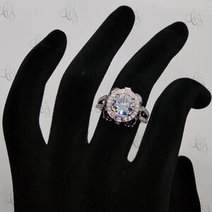 May include: A silver ring with a large, round, clear stone surrounded by smaller stones. The ring is on a black hand model.