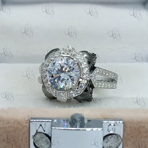 May include: A silver engagement ring with a large round center stone surrounded by smaller stones. The ring has a black halo around the center stone and a split shank with pave stones.
