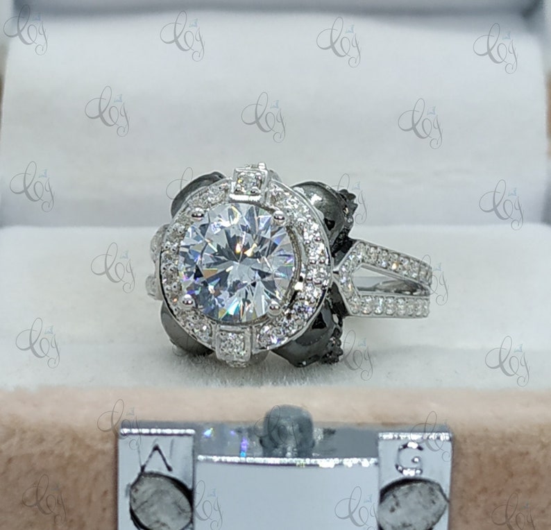 May include: A silver engagement ring with a large round center stone surrounded by smaller stones. The ring has a black halo around the center stone and a split shank with pave stones.