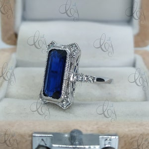 May include: A silver ring featuring a rectangular, deep blue gemstone. The ring has an ornate, filigree design around the stone and along the band, accented with small, clear stones. The ring is set in a light-colored jewelry box.