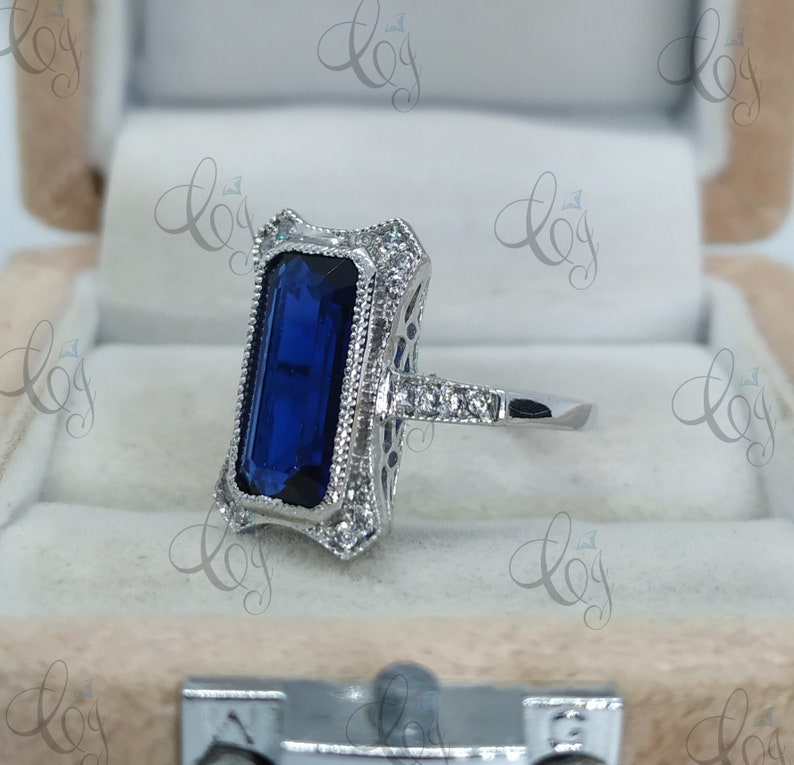 May include: A silver ring featuring a rectangular, deep blue gemstone. The ring has an ornate, filigree design around the stone and along the band, accented with small, clear stones. The ring is set in a light-colored jewelry box.