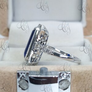 May include: A silver ring featuring a large, rectangular, dark blue gemstone. The gemstone is surrounded by a halo of smaller, clear stones and intricate filigree detailing. The ring's band is smooth and polished.