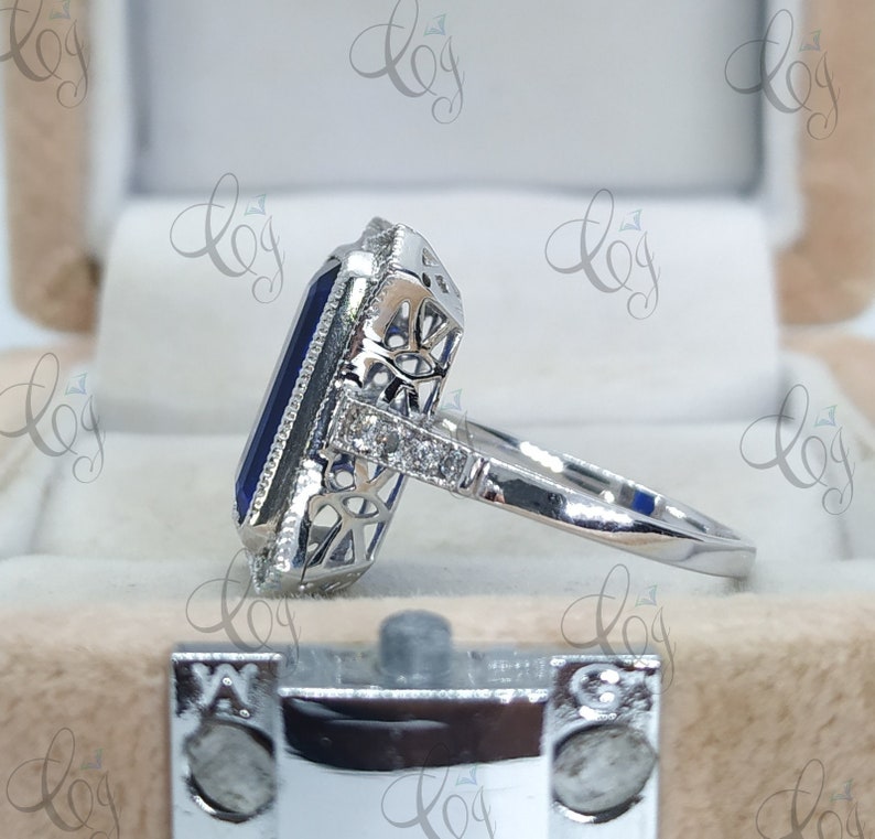 May include: A silver ring featuring a large, rectangular, dark blue gemstone. The gemstone is surrounded by a halo of smaller, clear stones and intricate filigree detailing. The ring's band is smooth and polished.