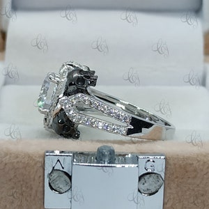 May include: A silver ring with a large, clear gemstone surrounded by smaller stones. The band of the ring features two black skull designs.