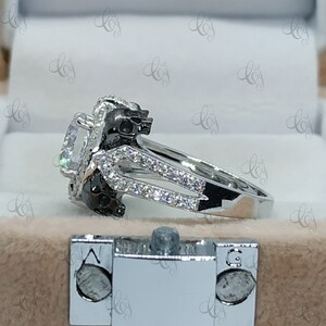 May include: A silver ring with a large, clear gemstone surrounded by smaller stones. The band of the ring features two black skull designs.