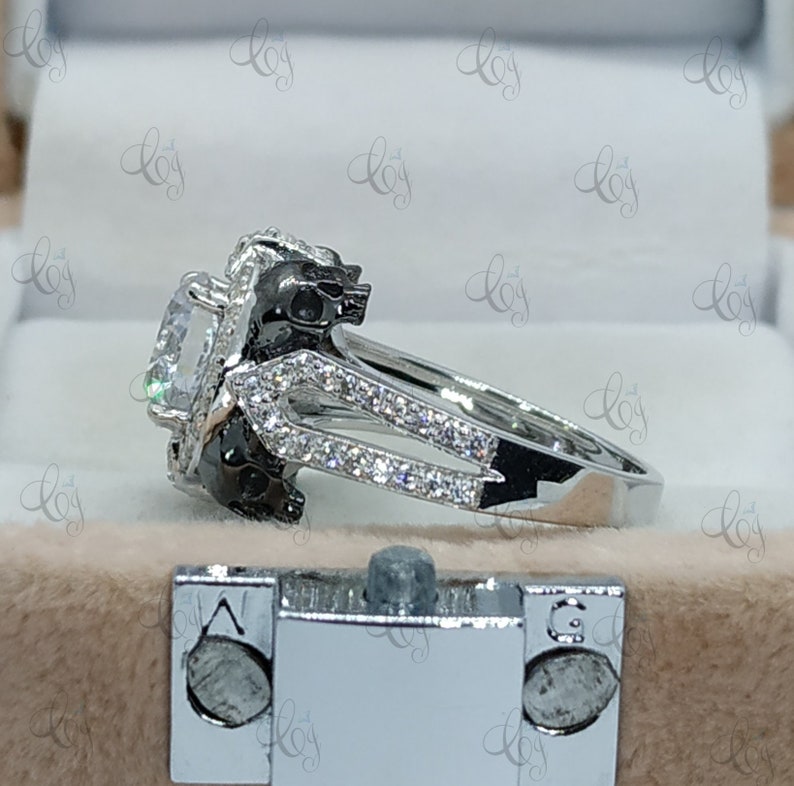 May include: A silver ring with a large, clear gemstone surrounded by smaller stones. The band of the ring features two black skull designs.