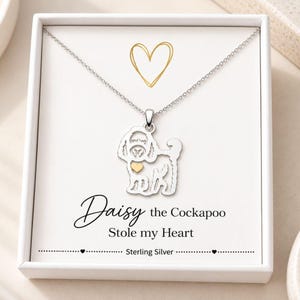 May include: A sterling silver necklace featuring a dog-shaped pendant with a small gold-coloured heart accent. The pendant hangs from a delicate chain inside a white gift box. The box insert displays a gold heart outline and the text Daisy the Cockapoo Stole my Heart.