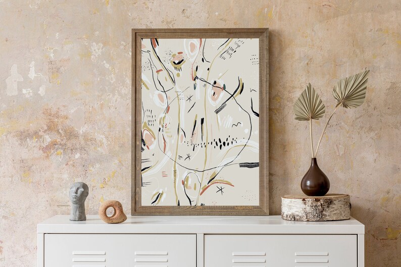 May include: A framed abstract art print with black, white, pink, and gold lines and shapes on a beige background. The print is hanging on a beige wall above a white dresser with a small grey sculpture and a wooden ring on top.