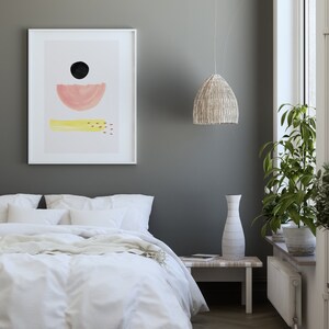 May include: A white framed print with a minimalist abstract design featuring a black circle, a pink half-circle, and a yellow rectangle with red dots. The print is hanging on a gray wall above a white bed with white bedding and a yellow throw blanket.