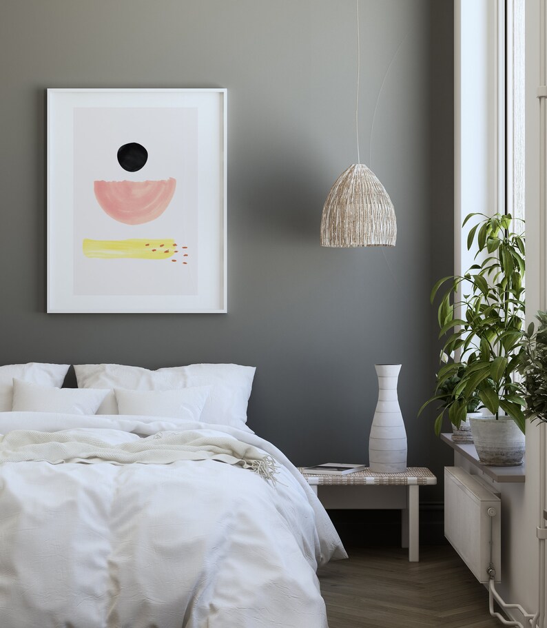 May include: A white framed print with a minimalist abstract design featuring a black circle, a pink half-circle, and a yellow rectangle with red dots. The print is hanging on a gray wall above a white bed with white bedding and a yellow throw blanket.