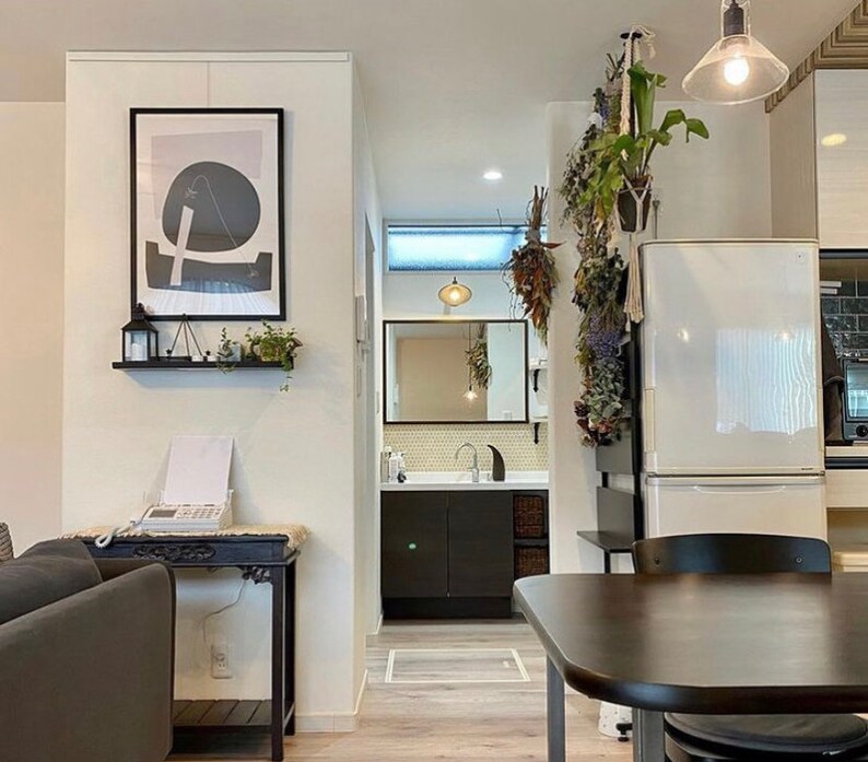 Puede incluir: Un refrigerador blanco con una mesa y sillas negras delante. Hay una estanter&iacute;a negra con una impresi&oacute;n abstracta en blanco y negro colgada en la pared sobre la mesa. Hay plantas colgando del techo sobre el refrigerador.