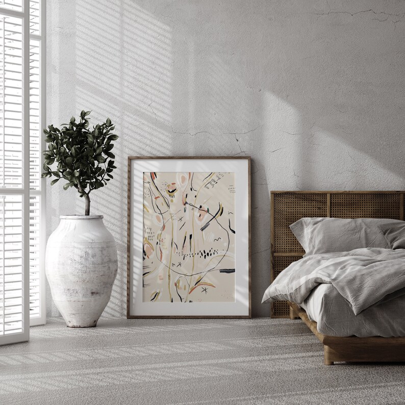 May include: A framed abstract art print with black lines and colorful shapes on a beige background. The print is hanging on a white wall in a room with a bed and a plant.