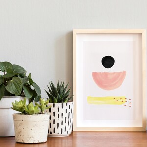 May include: A framed abstract print with a black circle, pink half-circle, and yellow stripe on a white background. Three potted plants are on a wooden surface in front of the print.